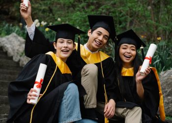 Three diverse graduates joyfully celebrating outdoors in caps, gowns, and holding diplomas.