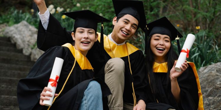 Three diverse graduates joyfully celebrating outdoors in caps, gowns, and holding diplomas.