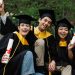 Three diverse graduates joyfully celebrating outdoors in caps, gowns, and holding diplomas.