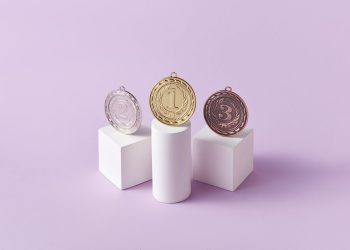 Gold, silver, and bronze medals displayed on white pedestals with a pink background.