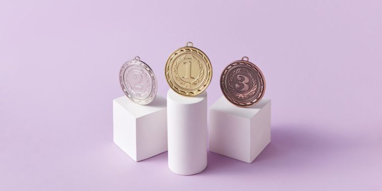 Gold, silver, and bronze medals displayed on white pedestals with a pink background.