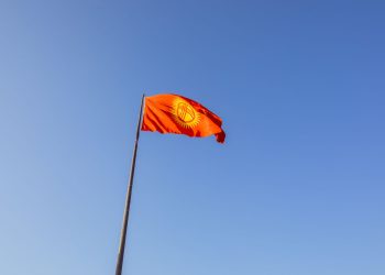 Red national flag of Kyrgyzstan waving on a flagpole under a clear blue sky.