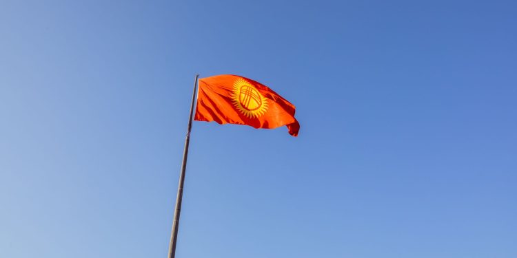 Red national flag of Kyrgyzstan waving on a flagpole under a clear blue sky.
