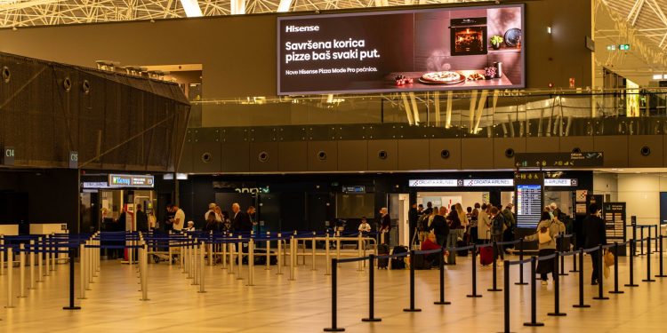 Travelers check in at Zagreb Airport with large advertising screen above.
