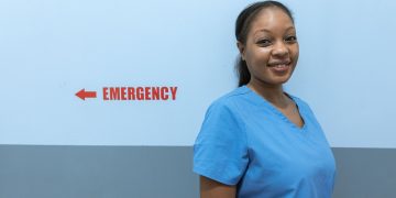 Happy healthcare worker in scrubs standing near emergency room sign.