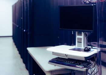 Contemporary computer with black screen placed on stand near row of server steel racks in data center