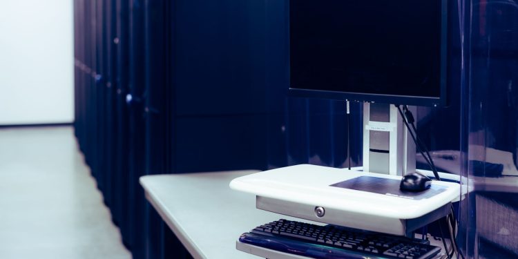 Contemporary computer with black screen placed on stand near row of server steel racks in data center