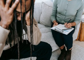 Crop concerned African American male sitting on sofa with female psychologist and holding head in hands in dismay during psychotherapy session