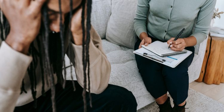 Crop concerned African American male sitting on sofa with female psychologist and holding head in hands in dismay during psychotherapy session