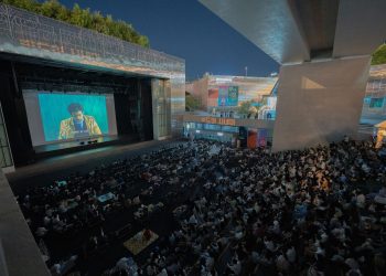 A large audience gathers outdoors at night to watch a movie screening under a bridge.