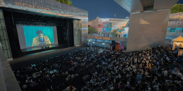 A large audience gathers outdoors at night to watch a movie screening under a bridge.