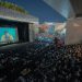 A large audience gathers outdoors at night to watch a movie screening under a bridge.