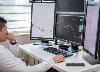 Business professional analyzing financial data on multiple computer monitors at his workspace.