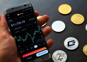 Close-up of a smartphone app showing Bitcoin trading details with crypto coins on a black surface.