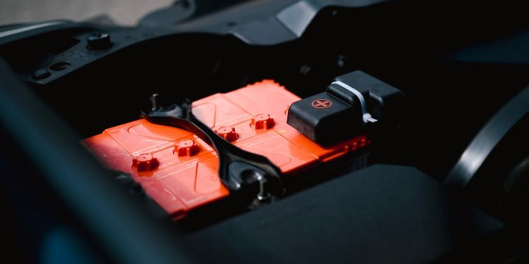Detailed view of an orange car battery inside a vehicle's engine bay, highlighting its features.