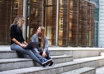 Two women sitting on steps with a laptop, discussing ideas in urban setting.