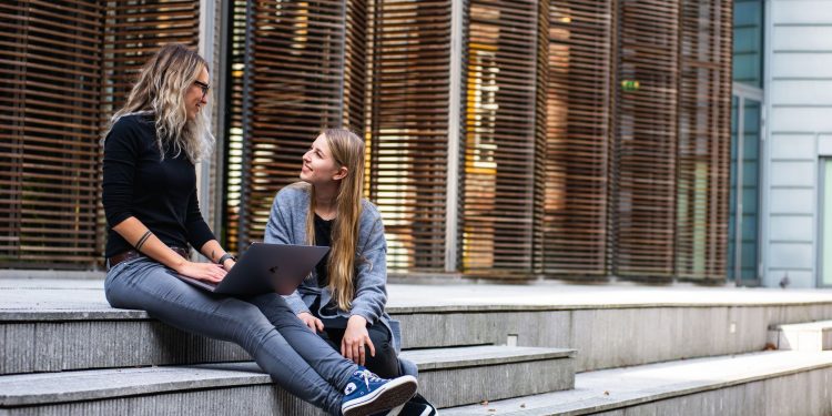 Two women sitting on steps with a laptop, discussing ideas in urban setting.