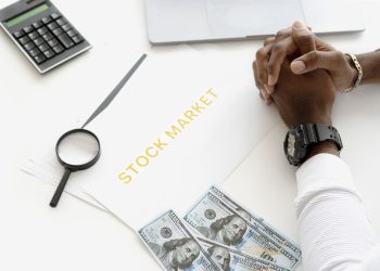 Hands folded over stock market documents with calculator and cash on desk.