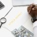 Hands folded over stock market documents with calculator and cash on desk.