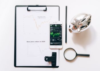 Top view of financial analysis tools including a cellphone, clipboard, magnifying glass, and piggy bank on a white desk.