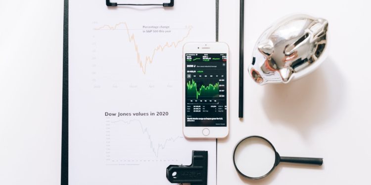 Top view of financial analysis tools including a cellphone, clipboard, magnifying glass, and piggy bank on a white desk.