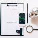 Top view of financial analysis tools including a cellphone, clipboard, magnifying glass, and piggy bank on a white desk.