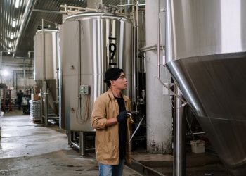 Asian man inspecting stainless steel tanks in a brewery, showcasing modern industrial processes.