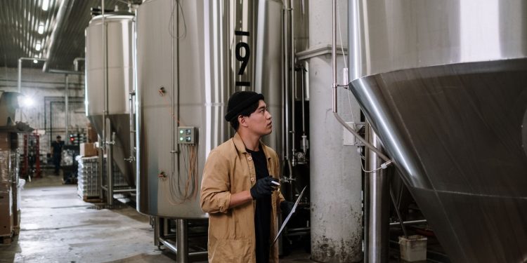 Asian man inspecting stainless steel tanks in a brewery, showcasing modern industrial processes.