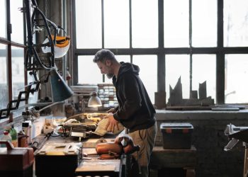 Side view of male workshop owner standing near messy workbench and fixing metal details while using instruments during work