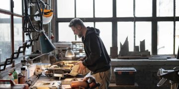 Side view of male workshop owner standing near messy workbench and fixing metal details while using instruments during work