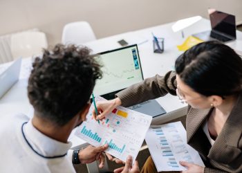Two colleagues in a meeting room discussing financial charts and graphs on a laptop and paper.