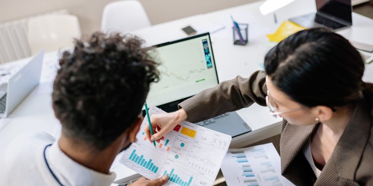 Two colleagues in a meeting room discussing financial charts and graphs on a laptop and paper.