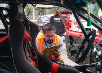 A man inspects a race car interior at a motorsport event, noting findings on a clipboard.