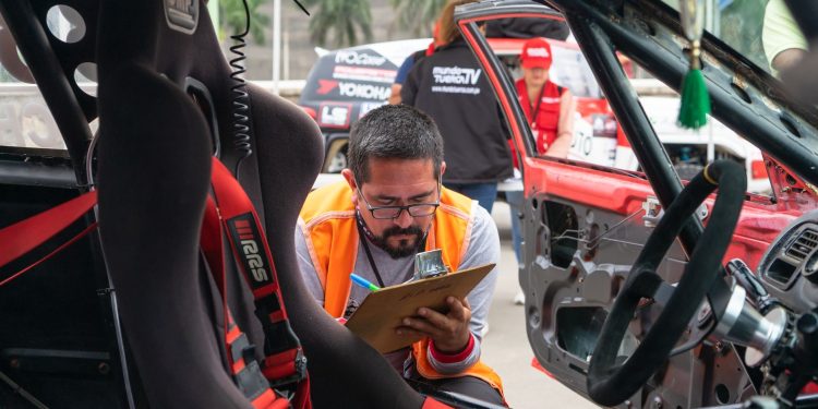 A man inspects a race car interior at a motorsport event, noting findings on a clipboard.