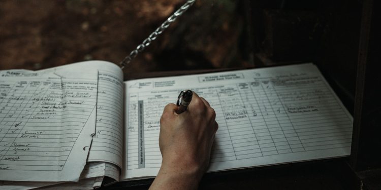 Close-up of a person writing in an outdoor logbook on a table.