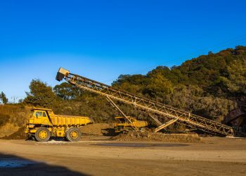 Yellow dump trucks and conveyor machinery in a sunny mining site with clear blue sky.