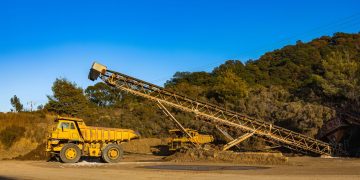 Yellow dump trucks and conveyor machinery in a sunny mining site with clear blue sky.