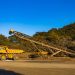 Yellow dump trucks and conveyor machinery in a sunny mining site with clear blue sky.