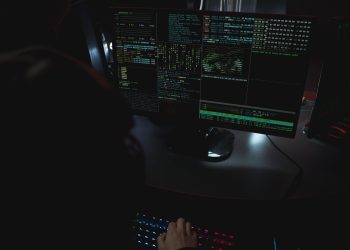 Dark room setup with code displayed on PC monitors highlighting cybersecurity themes.
