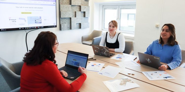 Three women collaborating at a conference table in a modern office with laptops and presentation screen.