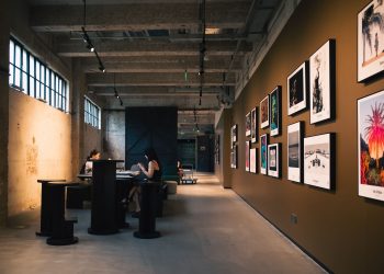 Art gallery hallway with framed artworks and visitors seated inside, under warm lighting.