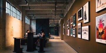 Art gallery hallway with framed artworks and visitors seated inside, under warm lighting.