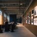 Art gallery hallway with framed artworks and visitors seated inside, under warm lighting.