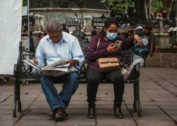 A man reads a newspaper and a woman uses her phone on a park bench in an urban setting.