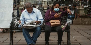 A man reads a newspaper and a woman uses her phone on a park bench in an urban setting.