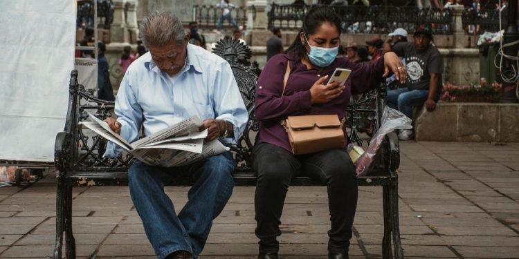 A man reads a newspaper and a woman uses her phone on a park bench in an urban setting.
