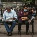 A man reads a newspaper and a woman uses her phone on a park bench in an urban setting.