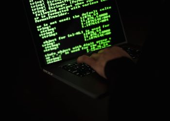 Crop unrecognizable computer geek typing on netbook with codes on screen while hacking system in darkness