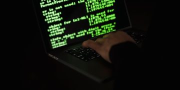 Crop unrecognizable computer geek typing on netbook with codes on screen while hacking system in darkness