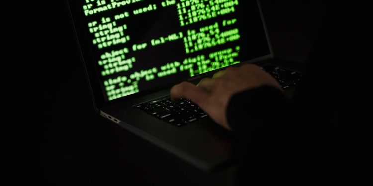 Crop unrecognizable computer geek typing on netbook with codes on screen while hacking system in darkness